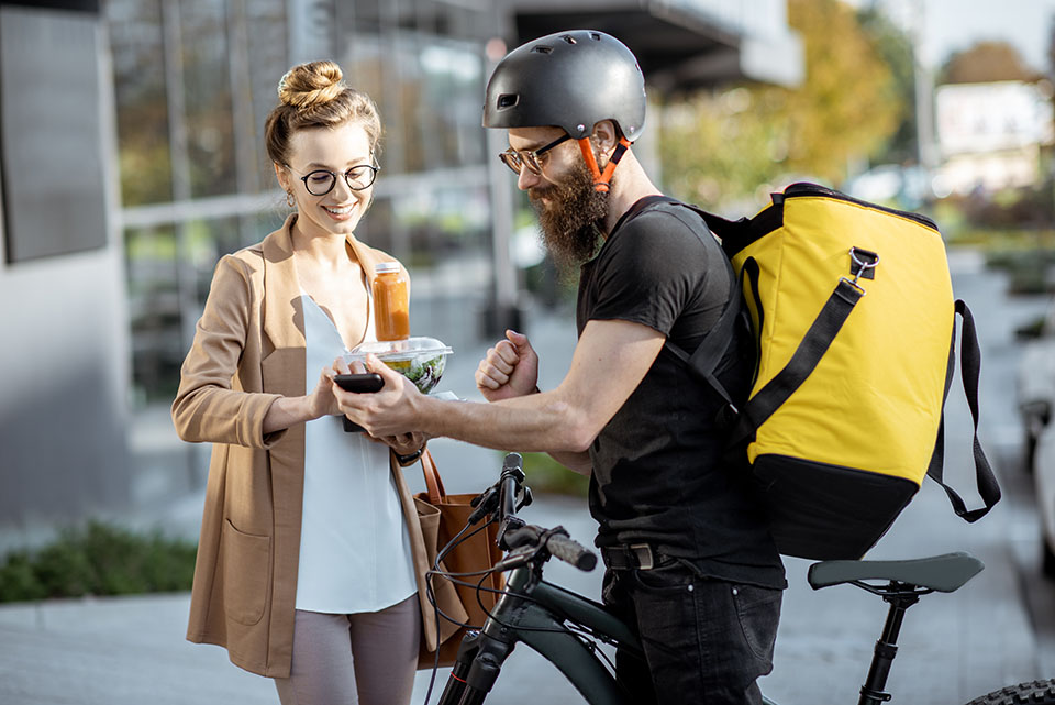 Gig Economy Courier delivering fresh lunches to a young business woman on a bicycle with thermal backpack. Takeaway restaurant food delivery concept